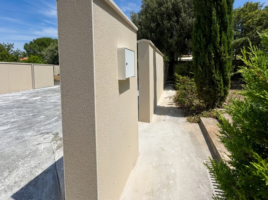 Entrée extérieure d'une propriété résidentielle, allée bétonnée longeant un mur en enduit taloché ton beige/sable