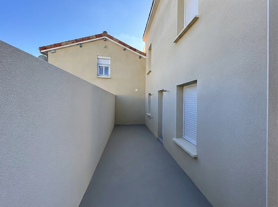 Passage extérieur étroit entre deux bâtiments résidentiels, délimité par un mur de clôture et une façade