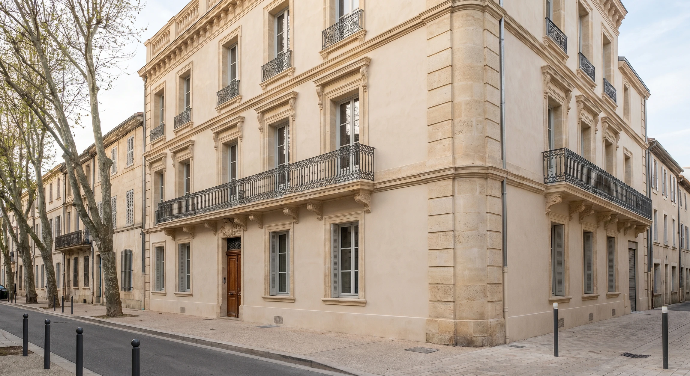 Photographie d'une façade d'immeuble ancien en pierre de taille (bâtiment de 3-4 étages, architecture de type belle époque ou fin XIXe), situé en milieu urbain ou semi-urbain. La façade bénéficie d'une réhabilitation visuelle manifeste : enduit rénové d'une teinte douce et homogène, moulures et éléments architecturaux soulignés et restaurés, fenêtres en bon état, façade propre et bien entretenue mettant en valeur le charme historique du bâtiment. Lumière naturelle douce mettant en valeur la texture et la qualité de l'enduit. Aucun ouvrier ni échafaudage visible. Photo prise de face ou légèrement en angle, de qualité professionnelle.