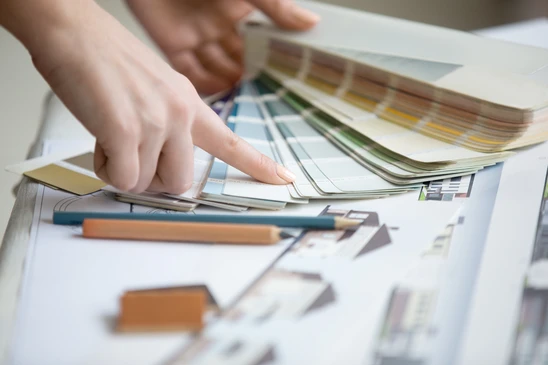 Personne consultant des nuanciers de couleurs et échantillons de matériaux sur un bureau de travail avec plans architecturaux et crayons de couleur
